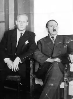 Achromatic, slightly torn photo print of two suited men indoors: to the left sits a balding blond eying the camera (with a rolled object in his breast pocket) and to the right, a sunken-jowled Hitler.