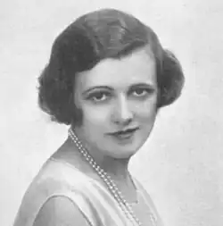Young white woman with dark hair in 1920s day clothes and a pearl necklace, looking towards the camera