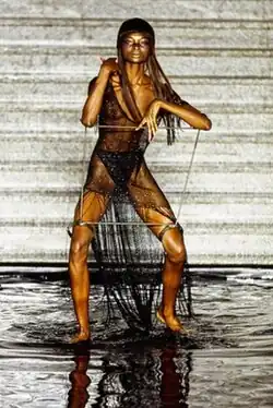 A Black woman wearing a sheer black dress, her elbows and thighs locked to a square metal frame