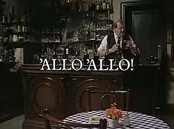 A man in a white shirt and dark vest stands behind a wooden bar, with bottles on shelves in the background. The words 'ALLO 'ALLO!' are superimposed in stylized white text across the center of the image