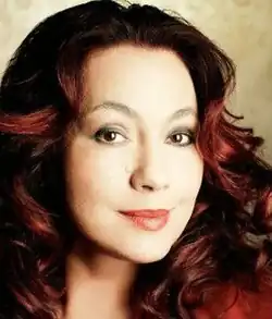Colour, three-quarter profile, studio, glamour, publicity portrait, of a smiling young woman wearing makeup. She has long, auburn wavy hair.