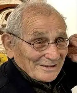 Three-quarter profile, colour Press shot, of a smiling, bespectacled, clean-shaven, elderly man, with thinning grey hair