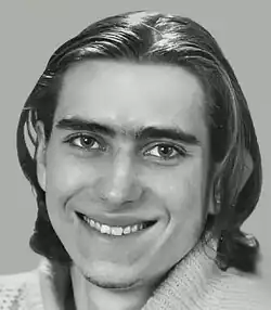 Black and white studio publicity headshot, of a smiling, clean-shaven man. He has shoulder-length, dark, straight hair with a central parting.