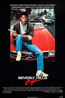 Axel Foley (portrayed by Eddie Murphy) sits on the hood of a red Mercedes-Benz convertible with a pistol pointing out on his left hand and his left foot resting above the word "Hills". The caption above reads "He is/has been chased, thrown through a window, and arrested. Eddie Murphy is a Detroit cop on vacation in Beverly Hills."