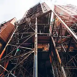 Low-angle photo of an urban apartment building. In the foreground, an overhanging portion is supported by rusty scaffolding.