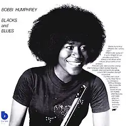 A black-and-white photo of the artist sat smiling and looking off to the side while holding a flute.