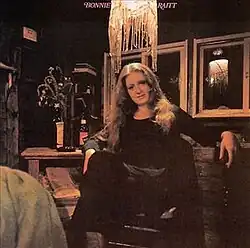 photo of Bonnie Raitt in black seated in interior scene under hanging lamp with table and bottles