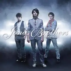The band members wearing suits, standing in a triangular position on a gray scale background. The band's logo and song name appear at the center of the imagine.