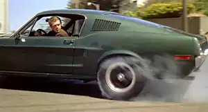Photograph of a car with a driver looking backwards out of its window: The car's rear tire is smoking from the friction of spinning against the road.