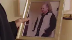 A man holds up a framed picture of a middle-aged man posing in a black and white shirt.