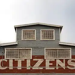 The cover art features a photo of a paint-chipped clapboard facade below a pale, overcast sky. The two-story facade is light teal and holds four faded industrial windows (the ground floor windows are double-width, whereas the first floor windows are single-width). On the ground floor, overlapping downwards from the lower-third of the windows, is found a wide red band, holding the block-lettered eponymous album title in cream, matching the roof fascia and industrial window trim.