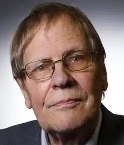 Colour publicity shot of a half-smiling, clean-shaven, bespectacled man, with short, greying hair, brushed forward. he is wearing a suit and shirt.