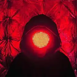 A person in a hoodie with a mask covering their face containing bright red lights arranged in a circle.