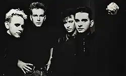 A group of young caucasion men, shot in black and white, looking at the camera. The man on th eleft has blonde hair, the rest have dark hair. They are all dressed in dark clothes and are shot from the waist up in a dark room.