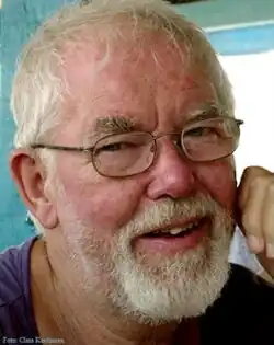 Colour three-quarter profile Press shot of a smiling, bespectacled man with white hair and full beard. He is wearing a tee shirt and resting his cheek on his hand.