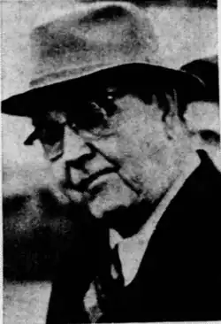 Candid black-and-white photo of an older Caucasian male wearing an overcoat, white shirt, tie, hat, and glasses