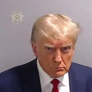Man wearing a dark blue suit, white shirt, and red tie. He stares into the camera lens from up close, with his chin tucked in, his lips pressed together, his forehead furrowed, with bags under his eyes. The light falls on his face from above to his left, with the right side of his face in deep shadow and his comb-over gleaming platinum blonde and sticking out on the left side. In the upper left corner of the image is the logo of the Fulton County sheriff's office, Patrick Labat.