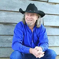 A picture of Doug Supernaw, seated and smiling while wearing a blue suit and cowboy hat