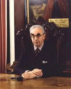 E. J. Kenney, an older white man with grey hair and bushy eyebrows, sitting in an ornate wooden chair in a formal hall, beneath a portrait of Dame Mary Ramsey. He wears a navy blazer, striped shirt, and regimental-style tie. A gavel lies on the table in front of him.