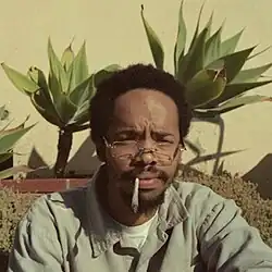 Earl is seen sitting and smoking a blunt of marijuana with two flowering plants and a brick wall behind him