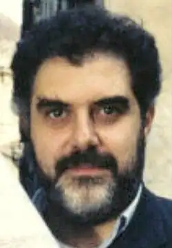 Colour Press shot of a bearded man with black, curly hair and full, trimmed beard which is slightly greying. He gazes to camera with striking, dark eyes and serious expression..