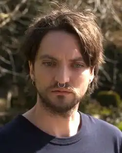 Erik (played by Richard Harmon) in a black shirt, resting his head on his right hand, looking into the camera