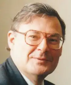 Colour publicity shot of a clean-shaven, smiling, bespectacled man, with short, greying, straight hair, parted on his left.