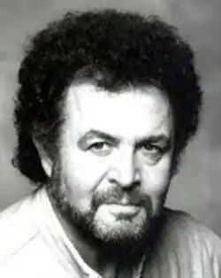 Black and white publicity headshot of a man with dark beard and moustache, curly dark hair in 1980s big-hair style, and dark eyes looking up at the camera from under dark brows.