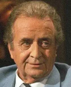 Colour publicity shot of a tanned, half-smiling, clean-shaven, elderly man in studio makeup and dyed hair, wearing a pale blue suit.