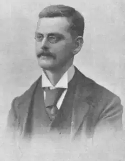 middle-aged white man with spectacles, neat moustache and short hair