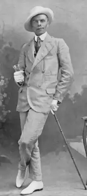 Young, clean-shaven white man in Edwardian day wear, wearing white gloves, white shoes and a white hat and leaning jauntily on a cane.