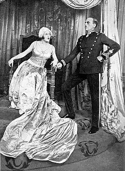 Young white woman seated on throne leaning provocatively towards an older white man standing stiffly at her side