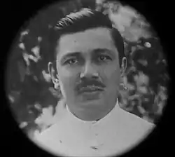 A black-and-white photograph of a mustached man in a white shirt with slicked-back hair.