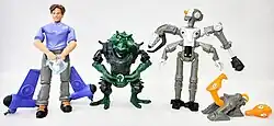 A photograph of four action figures on a white background. The figures are a boy wearing a blue shirt with rocket boosters on his legs; a green reptilian alien creature wearing black armor; a grey robotic figure with four arms and a single orange eye; and a small robotic figure with an orange alien head.