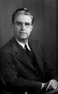 A middle-aged bespectacled man in a suit sitting with his hands crossed in his lap