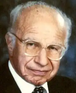 Three-quarter profile, colour publicity shot of an elderly, bespectacled, half-smiling man with short, receding white hair.