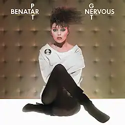photo scene of Pat Benatar with teased hair wearing a straightjacket looking very surprised, seated on the floor in a padded room
