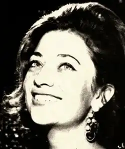 Three-quarter profile, black and white publicity shot of a smiling young woman with heavy makeup and long hair, under strong stage lighting.