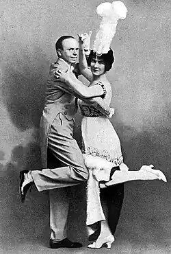 Young white couple in evening dress, dancing, both looking at the camera; she wears a tall, white-feather hat