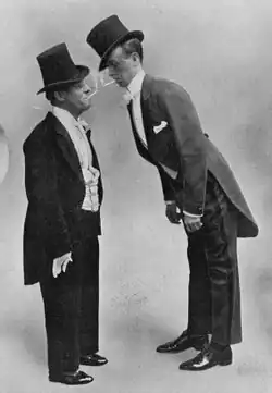 Short white man and tall white man standing in Edwardian evening costume wearing top hats and bowing to each other so that their lit cigarettes nearly meet.