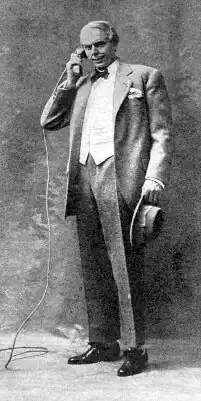 Young, clean-shaven white man in early 20th-century day wear, standing, holding his hat in one hand and talking into a telephone