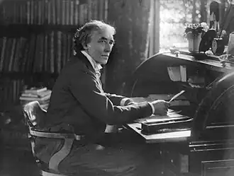 Middle-aged, clean-shaven white man with longish hair, seated at desk in booklined room, pen in hand