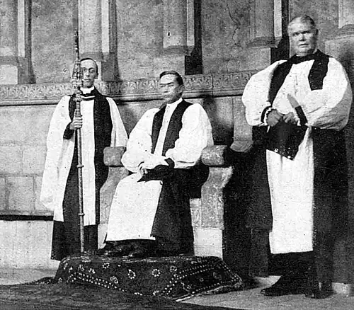 Interior of cathedral with clergyman robed and seated, flanked by an assistant carrying his crozier and another clergyman, standing, to his left