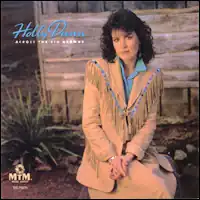 photo of Holly Dunn wearing sand colored western fringed jacket and skirt with blue blouse seated outside wood structure