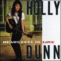 full-length photo of Holly Dunn wearing a white tee with black slacks strumming an acoustic guitar in front of windows of a structure