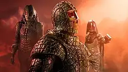 A trio of green, humanoid creatures. In the center is an armoured creature wearing a helmet with red eyes. Its jaw is visible, revealing a reptilian face and sharp teeth. To its left is a similar figure, though with its full body visible. It has similar armour, though has long dreadlocks of hair flowing behind it. To the right is another similar creature, except less armoured and with a different kind of helmet. It holds out its clawed hand as it looks to the camera.