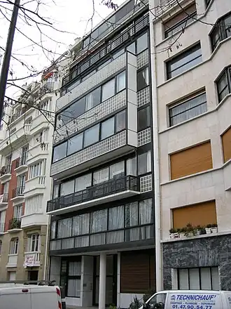 A modernist apartment with a facade of glass blocks, dark metal framing, and broad horizontal windows contrasts with the ornate, traditional buildings beside it