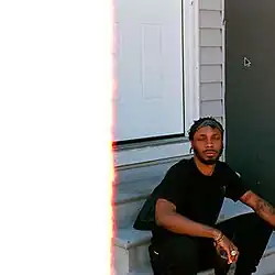 JPEGMafia sitting on a porch in front of a house with grey walls and a white door, he is holding a drink and his eyes are closed. Half of the photo is completely white due to a first of the (film) roll burn effect