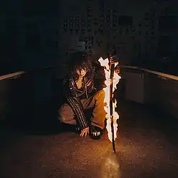 A person kneels down with a cigarette in their mouth, while pressing a burning katana onto the ground