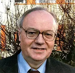 A balding man in a suit and square glasses with thick rims